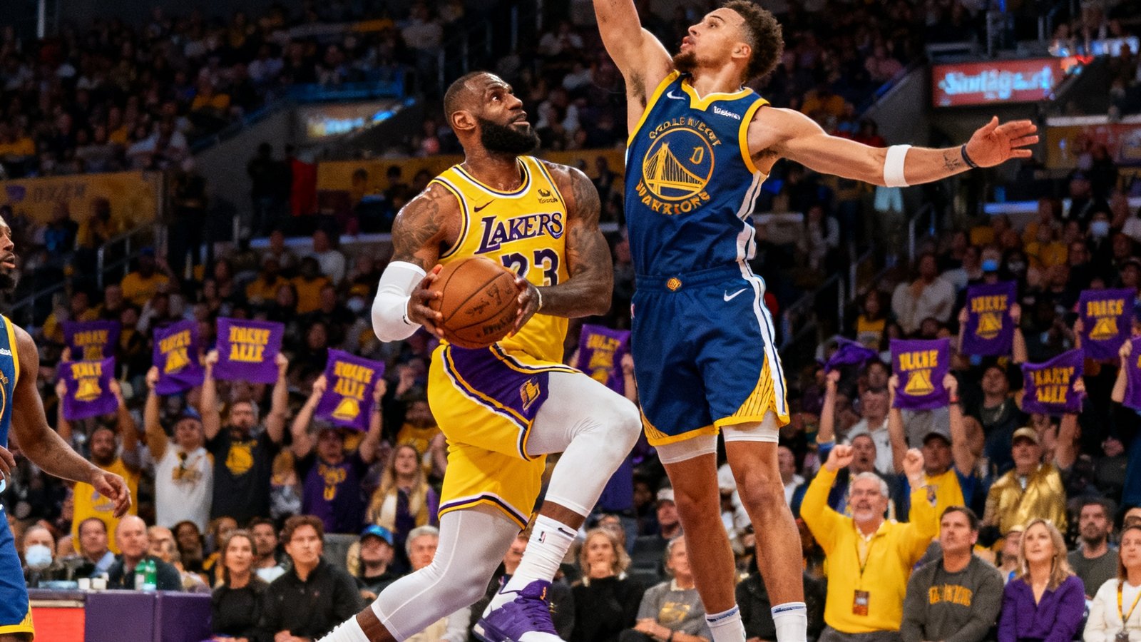 NBA战报配图：湖人主场击败勇士，詹姆斯持球突破上篮，防守球员试图封盖，斯台普斯中心观众挥舞紫金毛巾。
